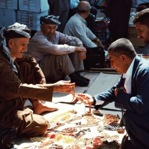 Elderly street vendor selling necklaces in a bustling outdoor market.