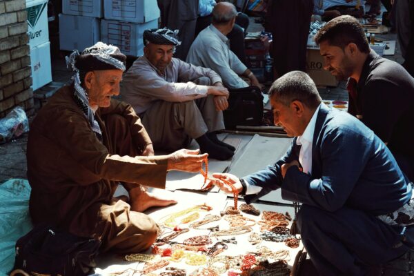 Elderly street vendor selling necklaces in a bustling outdoor market.