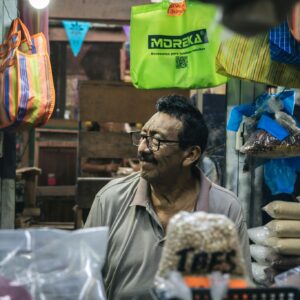 Friendly vendor in a local marketplace surrounded by dry goods, bags, and colorful signage.