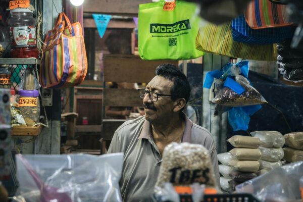 Friendly vendor in a local marketplace surrounded by dry goods, bags, and colorful signage.